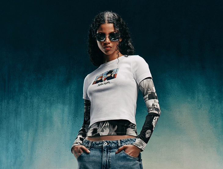 Woman wearing sunglasses and a white T-shirt over printed sleeves stands with hands in her pockets against a dark background. (photo)